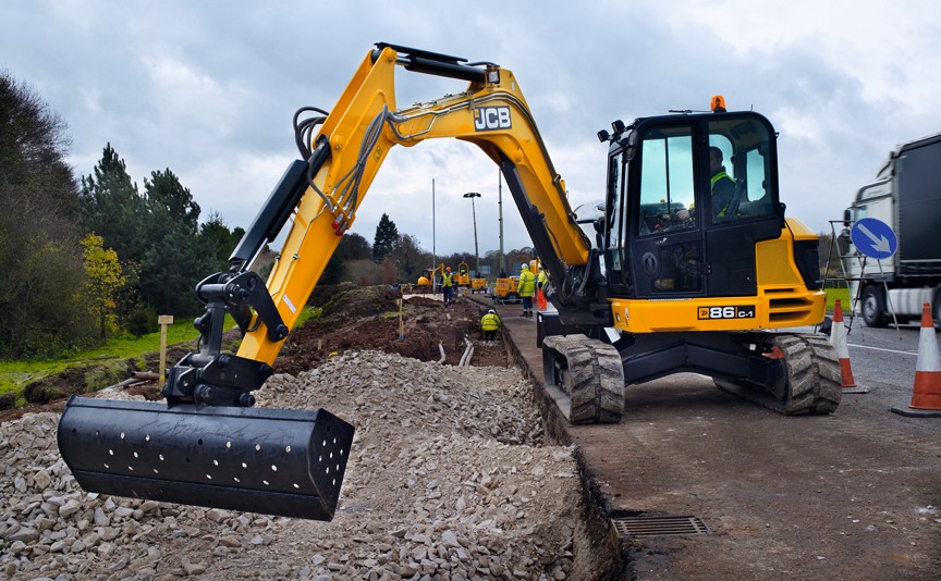 JCB compact excavator fitted with grading bucket, working on roadside construction with trenching and ground preparation taking place alongside traffic management cones.