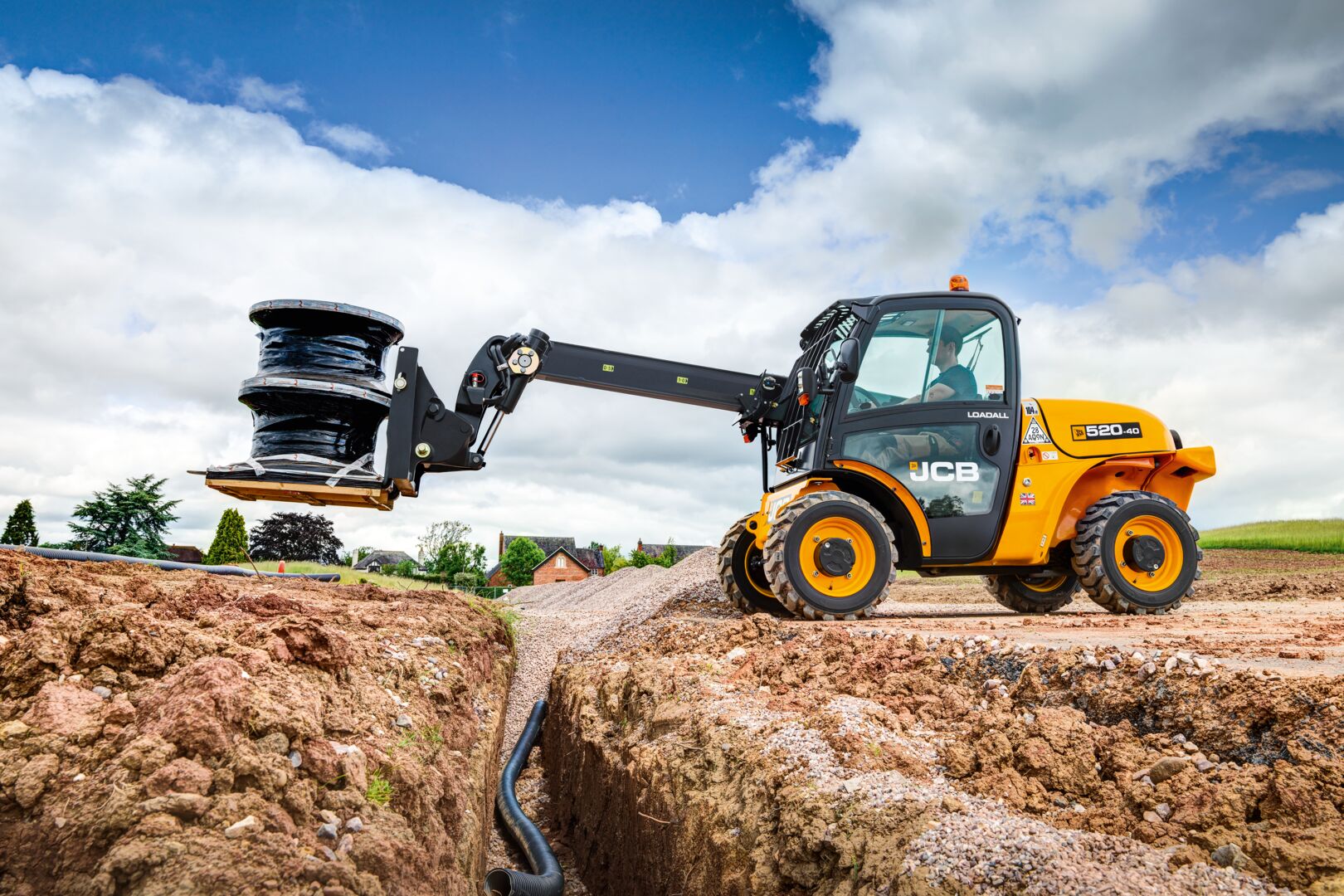 JCB 520-40 Loadall telehandler using fork attachment to lift a large spool of cable across a construction site trench, with exposed pipework, earthworks, and a cloudy sky in the background.