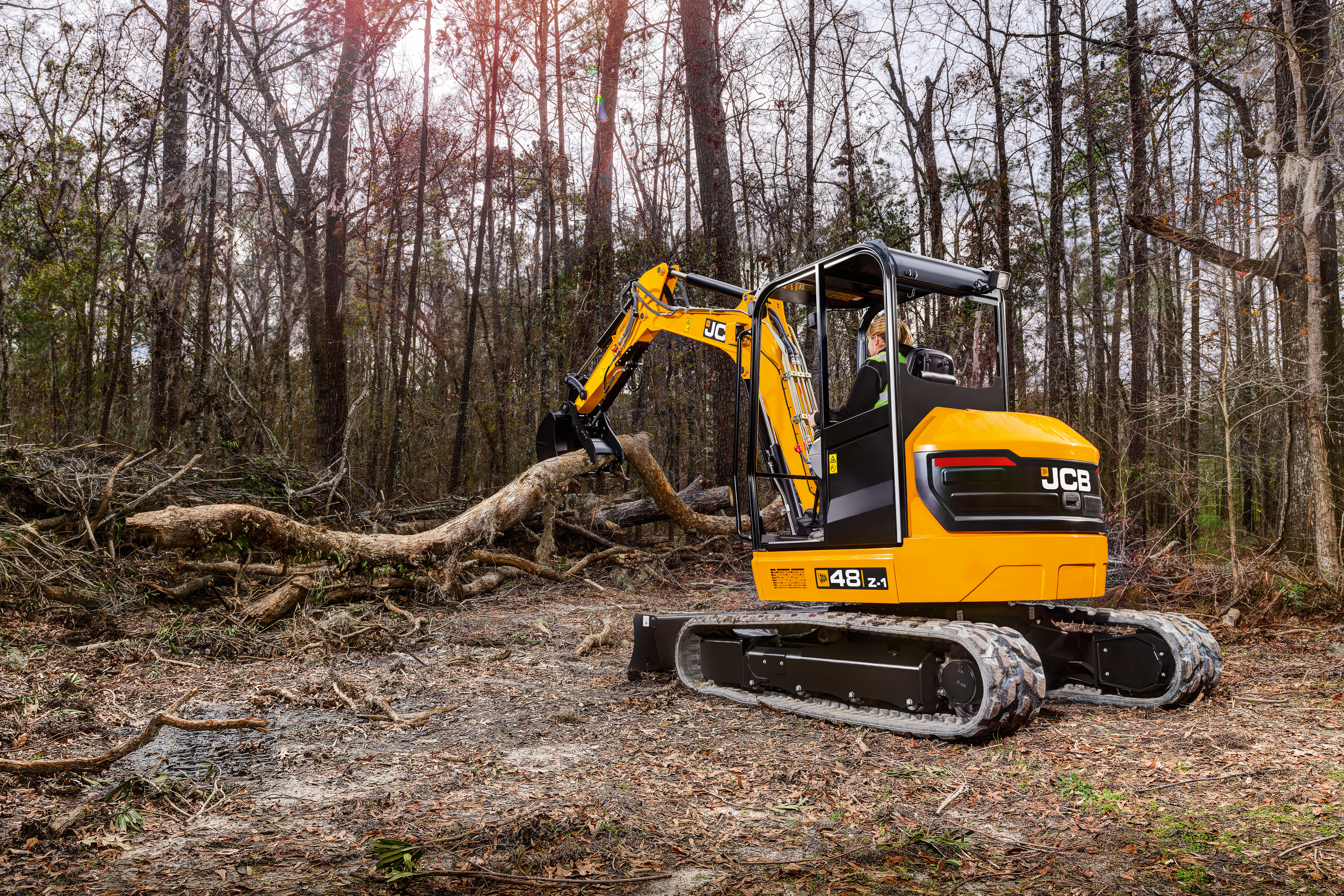 JCB 48Z-1 compact tracked excavator lifting a fallen tree trunk in a woodland clearing, featuring rubber tracks, enclosed cab and hydraulic boom during forestry and land clearance work