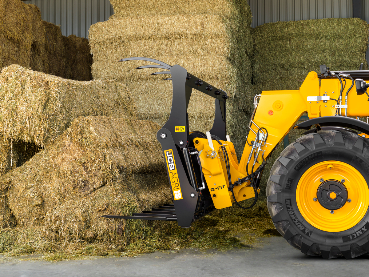 JCB Agri wheel loader fitted with a bale grab attachment handling rectangular hay bales inside a farm storage building, with stacked straw and silage bales arranged in the background.