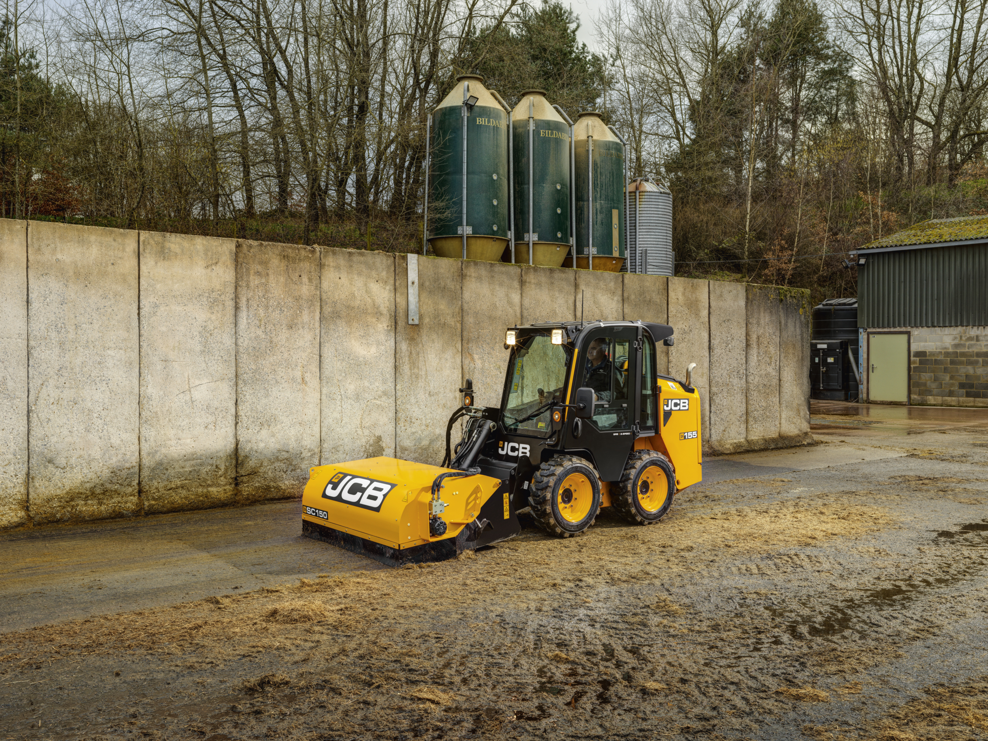 JCB 155 skid steer loader with SC150 sweeper attachment cleaning wet mud and straw on a farmyard, with concrete walls, feed silos, and trees in the background.