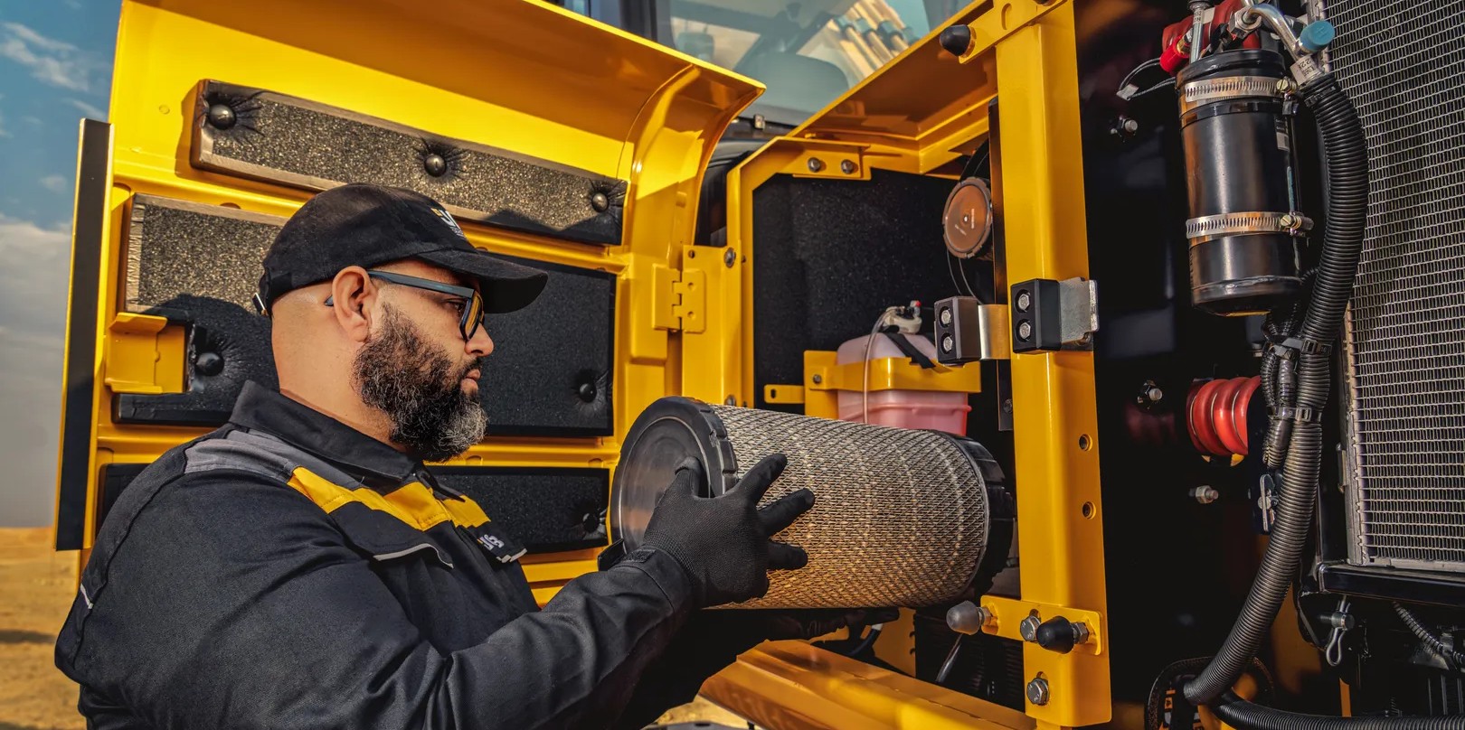Service technician replacing a cylindrical air filter inside the open engine compartment of a JCB excavator.
