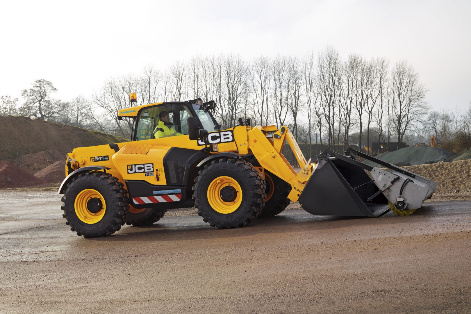JCB Loadall telescopic handler fitted with a bucket brush attachment, positioned on a construction site with aggregates in the background, demonstrating sweeping and material handling capabilities.