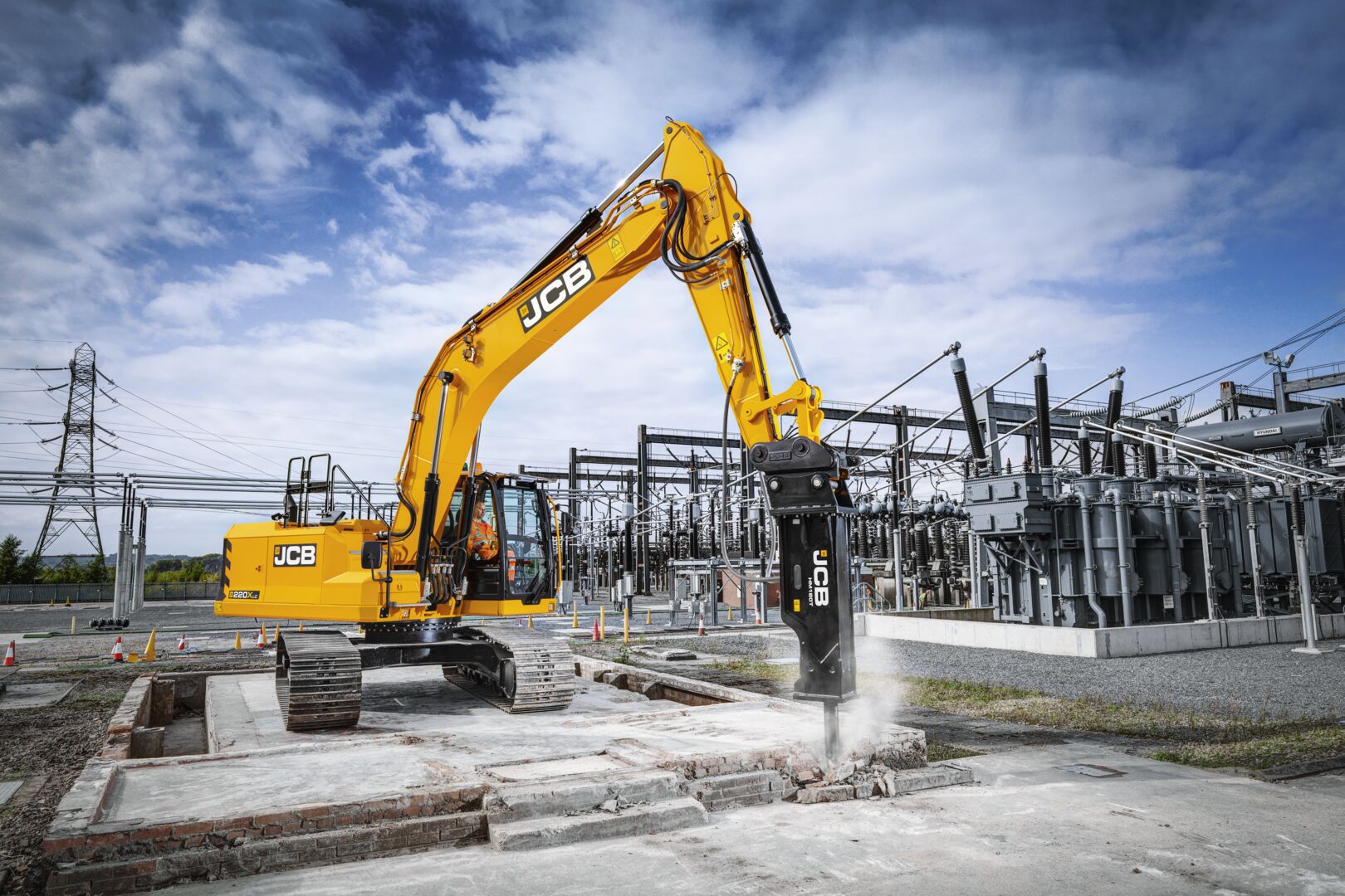 JCB tracked excavator fitted with hydraulic breaker attachment, breaking concrete at an industrial site with electrical substation infrastructure in the background.