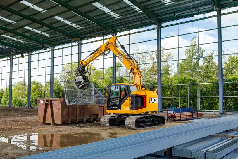 JCB tracked excavator fitted with a hydraulic grab attachment sorting and loading scrap metal inside a large industrial building, with steel beams, construction materials and floor-to-ceiling windows visible in the background.
