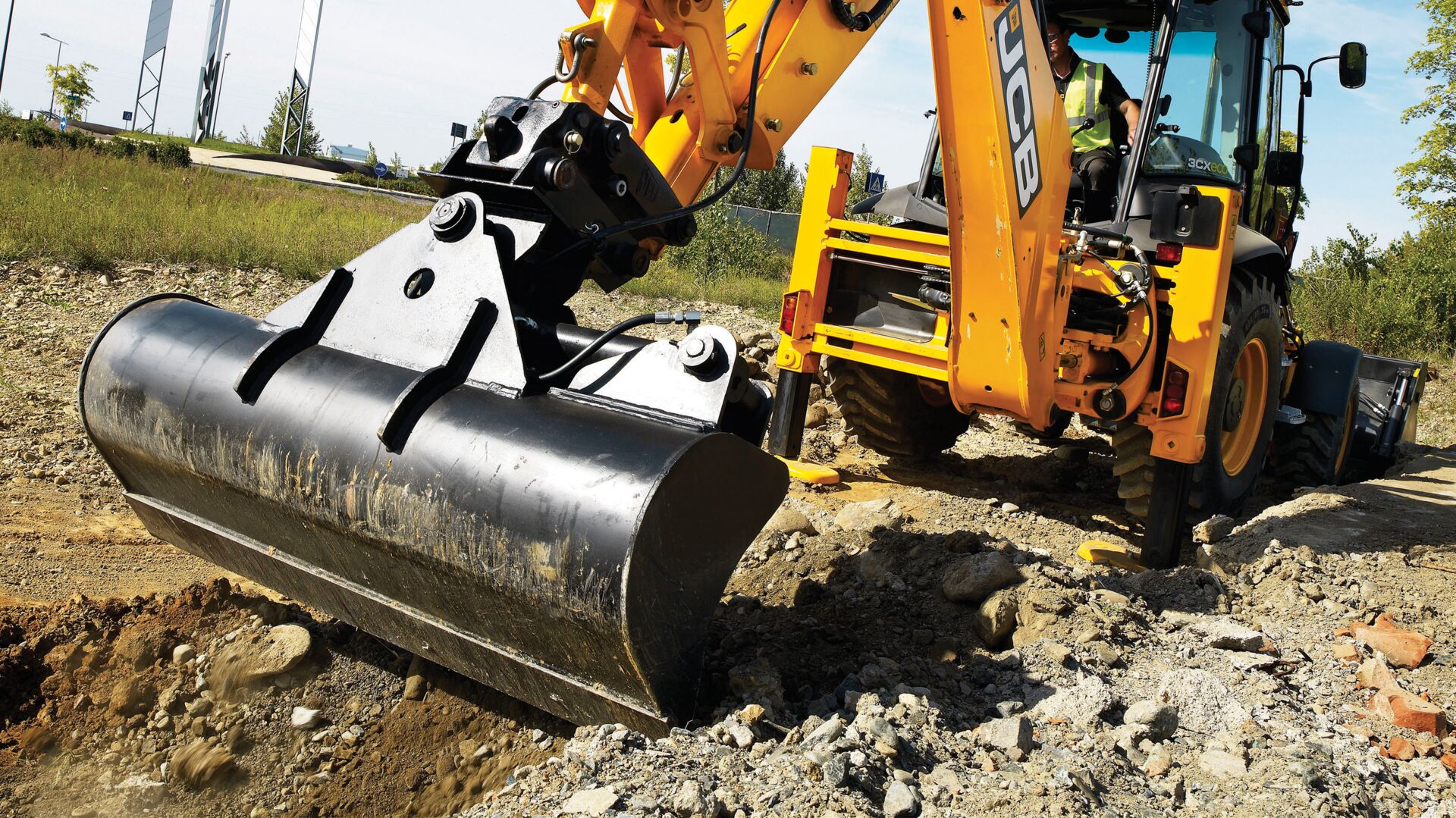 JCB tilting bucket attachment mounted on a JCB backhoe loader, grading and levelling rocky ground on a construction site. The hydraulic tilt mechanism allows precise bucket positioning for shaping surfaces and working on uneven terrain.