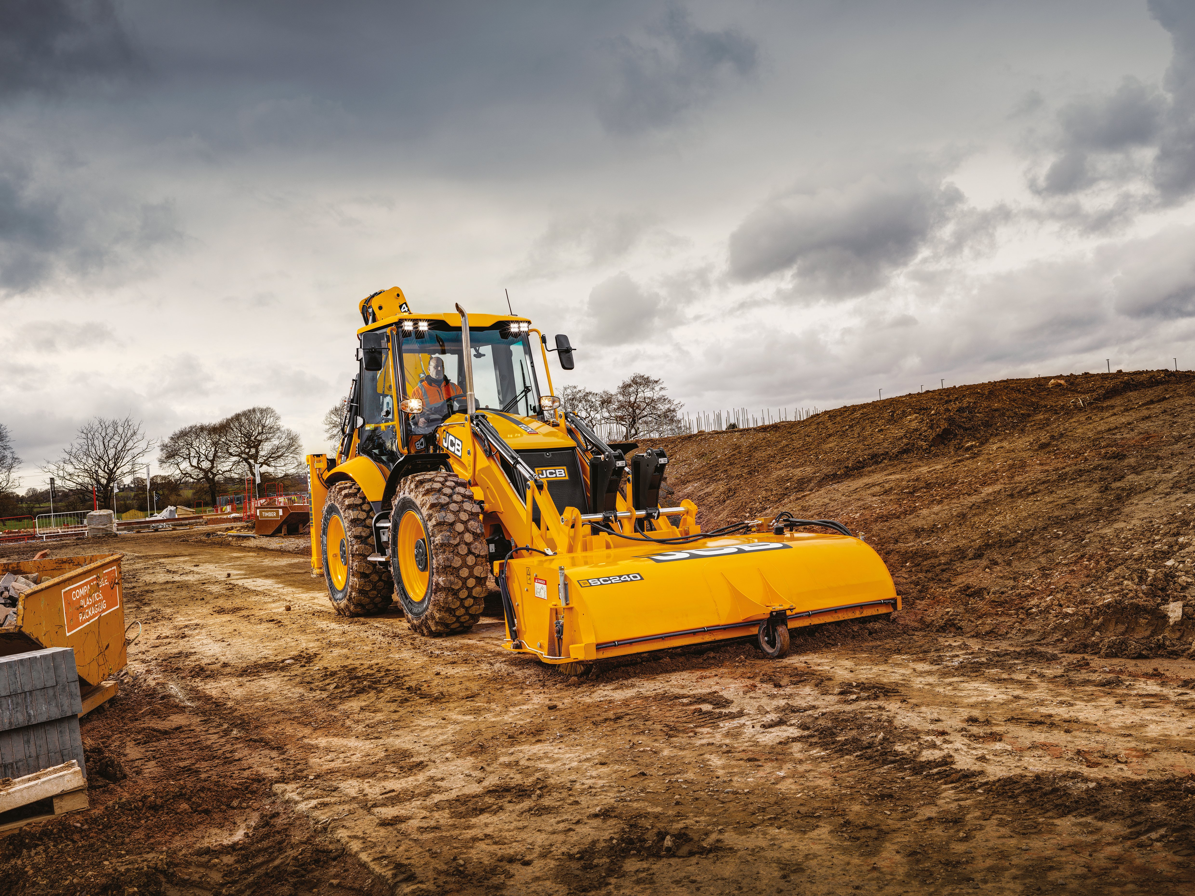 JCB backhoe loader fitted with an SC240 sweeper attachment operating on a muddy construction site under a cloudy sky, with a cab operator visible and building materials in the background.
