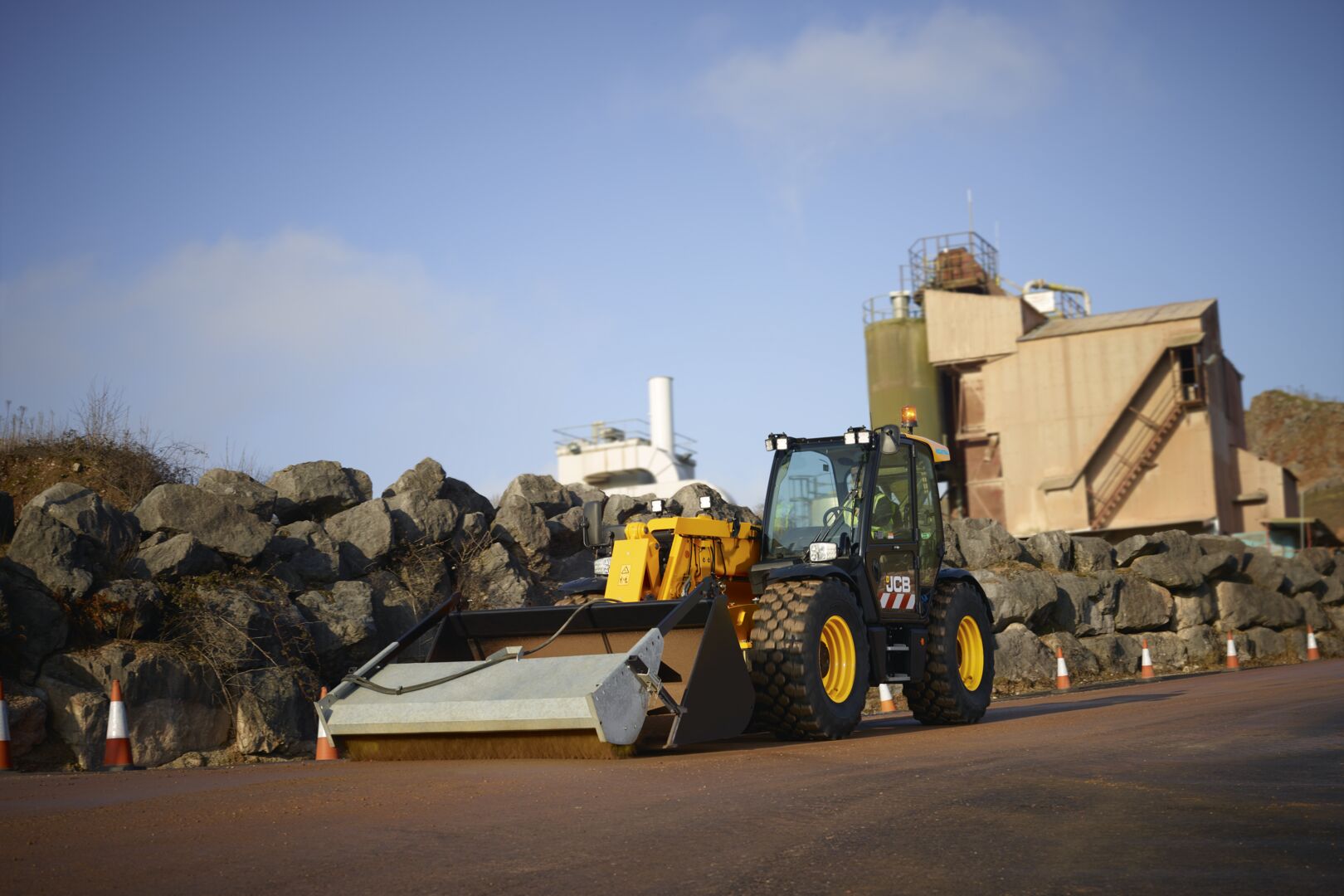 JCB telescopic handler fitted with a road sweeper attachment operating on a paved surface near an industrial quarry facility with rock embankments in the background.